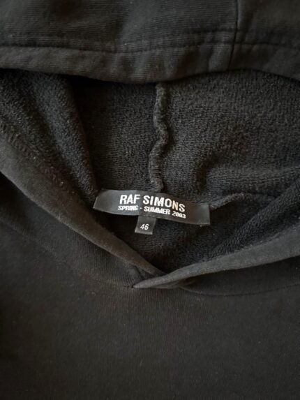 **Alt text:** Black Blood Vision graphic hoodie close-up showing the inner hood, drawstrings, textured fabric, and RAF SIMONS Spring-Summer 2026 label.